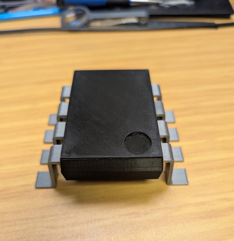 May include: A black electronic component with a rectangular body and grey metal pins. The component has a circular indentation on its top surface. The item is placed on a light brown wooden surface.