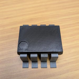 May include: A black electronic component with eight grey pins. The component is rectangular with a circular indentation on the top. The pins are evenly spaced and extend from the bottom.