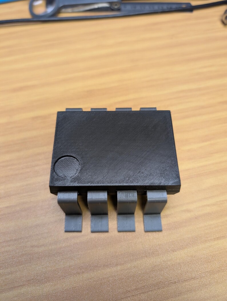 May include: A black electronic component with eight grey pins. The component is rectangular with a circular indentation on the top. The pins are evenly spaced and extend from the bottom.