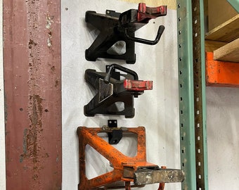 May include: Four orange and black car jacks are mounted on a white wall. The jacks are hanging from metal brackets. The jacks are in a vertical row, with the top jack being the oldest and the bottom jack being the newest. The jacks are all in good condition.