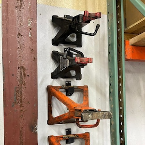 May include: Four orange and black car jacks are mounted on a white wall. The jacks are hanging from metal brackets. The jacks are in a vertical row, with the top jack being the oldest and the bottom jack being the newest. The jacks are all in good condition.