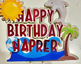 May include: A layered cake topper with the words "HAPPY BIRTHDAY HARPER" in red, with a yellow sun, a gray dolphin, a palm tree, and a blue wave design. The topper is on white sticks.