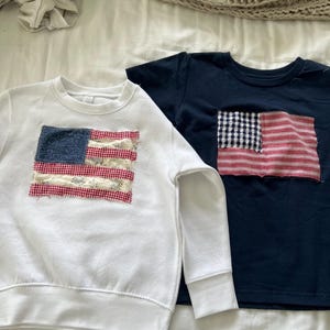 May include: Two children's shirts with American flag patches. One is a white sweatshirt, and the other is a navy blue t-shirt. The flags are made of patterned fabric in red, white, and blue.