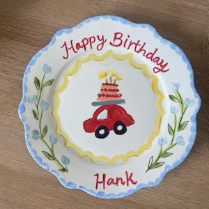 May include: A round ceramic plate with a scalloped edge painted in light blue. The center features a red car carrying a birthday cake, surrounded by a yellow sunburst. The text reads Happy Birthday at the top and the name Hank at the bottom.