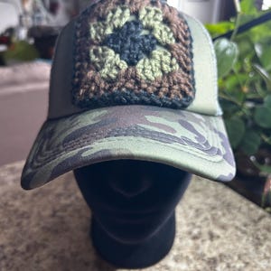 May include: A camouflage baseball cap with a crocheted square patch. The patch features a floral design in shades of green, brown, and black. The cap has a curved brim and a mesh back.
