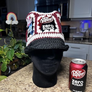 May include: A handmade crochet bucket hat with a Dr. Pepper Zero Sugar theme. The hat features a black brim, white and burgundy panels, and patches with the Dr. Pepper logo. A Dr. Pepper Zero Sugar can is next to the hat.