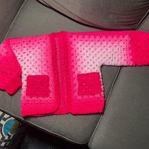 May include: A handmade, open-front crochet cardigan in shades of pink. The sweater features a gradient effect, transitioning from light pink to a vibrant fuchsia. It has two square pockets and long sleeves. The garment is displayed on a gray surface.
