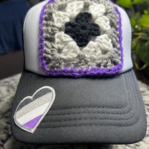 May include: A black and white trucker hat with a crocheted square patch in the center. The patch features a black cross design surrounded by white, gray, and purple crochet. A heart-shaped patch with gray, white, and purple stripes is on the brim.