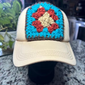 May include: A beige baseball cap featuring a square crochet patch on the front panel. The patch displays a central cream-colored square surrounded by rust-red and bright turquoise yarn in a classic granny square pattern with a dark brown border.