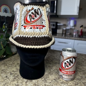 May include: A handmade crochet bucket hat featuring A&W Zero Sugar Root Beer can panels. The hat has a brown brim and a beige and white crochet design. A&W Zero Sugar Root Beer can is next to the hat.