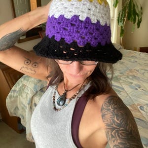 May include: A person wearing a crocheted bucket hat with yellow, white, purple, and black stripes. The hat is a representation of the non-binary pride flag.