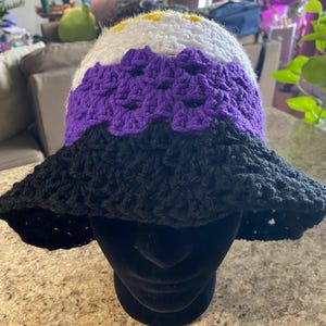 May include: A crocheted sun hat with a wide brim. The hat is black, purple, white, and yellow. The colors are arranged in stripes, with the black stripe at the bottom, followed by the purple stripe, then the white stripe, and finally the yellow stripe at the top.