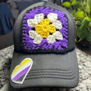 May include: A black trucker hat with a crocheted square featuring a yellow, white, and purple floral design. A non-binary heart-shaped patch is on the front. The hat has a curved brim and a mesh back.