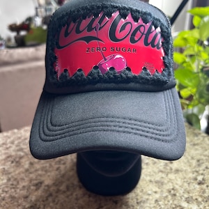 May include: Black trucker hat with a red Coca-Cola Zero Sugar logo featuring a cherry graphic. The hat has a black brim and a mesh back. The Coca-Cola logo is outlined with black fur.