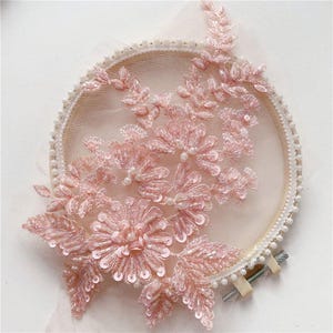 Puede incluir: Aplique de encaje floral rosa con lentejuelas y perlas sobre un fondo de malla blanca. El aplique es circular y tiene un diseño delicado y femenino.