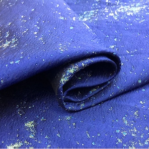 Op de afbeelding: Close-up van een blauwe stof met een metallic zilver en groen gevlekt patroon. De stof is over zichzelf gevouwen.