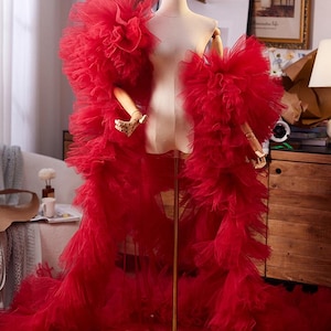 May include: A vibrant red floor-length robe crafted from layers of sheer tulle fabric. The garment features voluminous, ruffled sleeves and a full, flowing skirt that drapes down from a mannequin, creating a dramatic silhouette with dense texture.