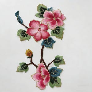 Puede incluir: Diseño floral bordado con flores rosas, ribetes dorados, hojas verdes y detalles azules. Las flores están en plena floración, con un patrón de puntadas detallado. El diseño está sobre un fondo blanco.