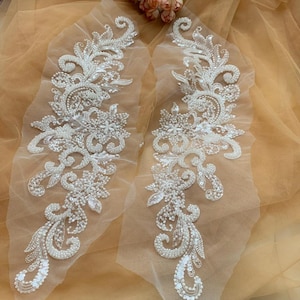 May include: Two white beaded lace appliques with pearl accents and a floral design. The appliques are on a sheer white mesh fabric.
