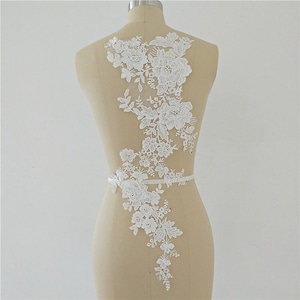 May include: A white floral lace applique, featuring a delicate arrangement of flowers and leaves. The applique is displayed on a mannequin, showcasing its design and potential use in bridal or formal wear. The lace is intricately detailed.