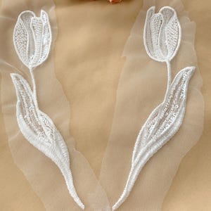 Könnte beinhalten: Zwei weiße, perlenbesetzte Tulpenblumen-Applikationen auf einem durchsichtigen weißen Netzstoff. Die Applikationen sind perfekt, um jedem Projekt einen Hauch von Eleganz zu verleihen.