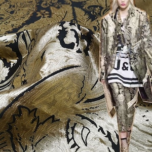 May include: Gold and black brocade fabric with a floral pattern. The fabric is shiny and has a textured surface. A person is wearing a jacket made of the same fabric.