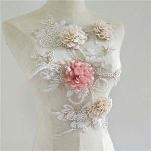 May include: A delicate lace applique with three-dimensional flowers. The applique features a pink flower, two cream-colored flowers, and intricate beading.