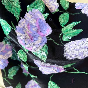 May include: Black fabric with large, sequined floral embroidery. The flowers are a gradient of purple and pink, with green sequined leaves. The fabric has a soft, velvety texture.