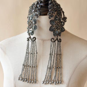 Puede incluir: Un collar adornado con cuentas grises y plateadas, con diseños florales y de hojas. El collar tiene largas hebras de cuentas colgantes, creando una estética vintage. El collar se exhibe en un maniquí.