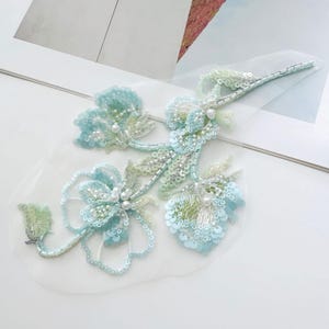May include: A floral applique patch featuring light blue and pale green sequins, glass beads, and small white faux pearls arranged in a botanical design on a sheer white mesh backing. The embroidery includes intricate beadwork and sequin detailing.