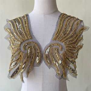 May include: Gold beaded and sequined fabric applique in the shape of wings. The applique is on a black mesh backing.
