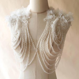 May include: A white beaded shoulder piece featuring cascading strands of clear and white beads. The design includes floral fabric appliqués on each shoulder, accented with small white pearls, creating a draped, layered effect across the chest.