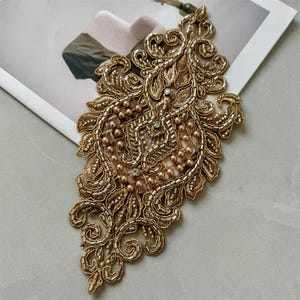 May include: An ornate gold embroidered applique featuring intricate scrollwork, metallic thread detailing, and small round beads. The decorative piece is embellished with tiny clear crystals scattered throughout the symmetrical floral-inspired pattern.