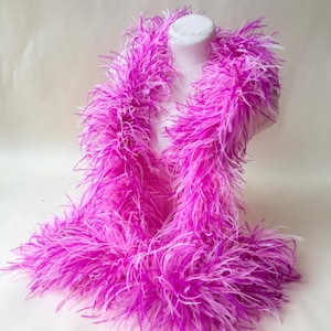 May include: A long, fluffy feather boa featuring a vibrant gradient of bright magenta and white feathers. The soft, textured accessory is draped around a plain, neutral-colored mannequin torso, showcasing the full volume and length of the plumage.