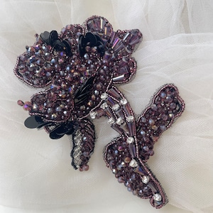 以下が含まれることがあります： 繊細なデザインの紫と黒のビーズでできた花柄のアップリケ。アップリケは、小さな虹色のビーズとスパンコールでできています。
