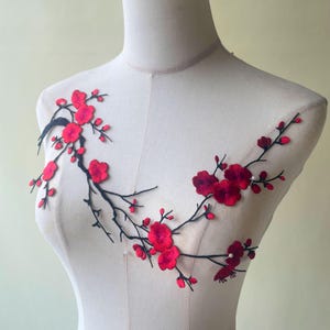 Könnte beinhalten: Gestickte Blumenapplikation mit einem Zweig mit roten Blüten und Knospen. Die Blüten haben ein geschichtetes Design und die Zweige sind schwarz. Die Applikation wird auf einer Schaufensterpuppe präsentiert.