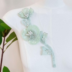 May include: A light mint green floral appliqué featuring intricate beadwork, sequins, and small white pearls. The design consists of two layered flower blossoms connected by a stem, all embellished with shimmering textures and delicate embroidery.