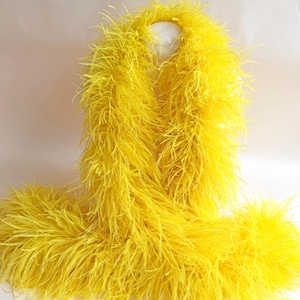 May include: A bright yellow feather boa made of long, wispy plumes. The accessory features a dense, voluminous texture with individual feathers extending outward, creating a soft and airy appearance throughout the entire length of the piece.