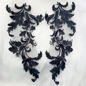 May include: Two black lace appliques with a floral design. The appliques are on a white background.