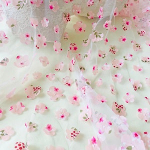 May include: White tulle fabric with pink 3D flower embellishments. The flowers have a pink center and green leaves.