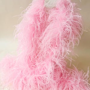 Pode incluir: Uma boá de penas longa e fofa em um tom suave de rosa pastel. O acessório é composto por inúmeras penas delicadas e finas que criam uma aparência volumosa e texturizada, caindo em uma forma solta e fluida com uma qualidade leve e arejada.