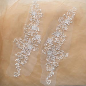 Puede incluir: Dos apliques de encaje floral blanco con intrincados diseños de flores y hojas. Los apliques están adornados con detalles brillantes y se colocan sobre un fondo de tela de color melocotón suave. Ideales para proyectos nupciales o de manualidades.
