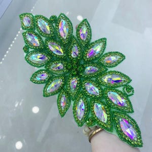 Puede incluir: Aplicación decorativa con diseño floral, con contornos de cuentas verdes y detalles de cristales iridiscentes. La aplicación mide aproximadamente 15 cm de largo y 7,5 cm de ancho, adecuada para adornar prendas o accesorios.