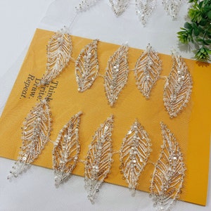 Pode incluir: Bordado de folhas brancas com contas em um tecido de malha branca. O bordado apresenta um padrão repetitivo de folhas com um design delicado e intrincado.