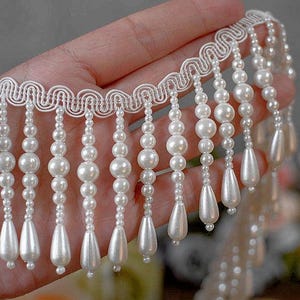 May include: White beaded fringe trim with a scalloped edge. The trim features white pearl beads of varying sizes, with teardrop-shaped beads at the bottom. This decorative trim is suitable for embellishing garments or crafts.