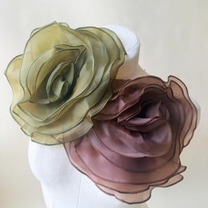 Op de afbeelding: Twee grote, gelaagde stoffen bloembroches bevestigd aan een witte etalagepop. De ene bloem is olijfgroen en de andere is gedempt mauvebruin. Beide bloemen hebben meerdere doorschijnende bloemblaadjes met afgewerkte, gestikte randen.