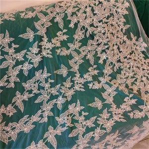 May include: White floral lace fabric with a green background. The lace has a delicate leaf pattern.