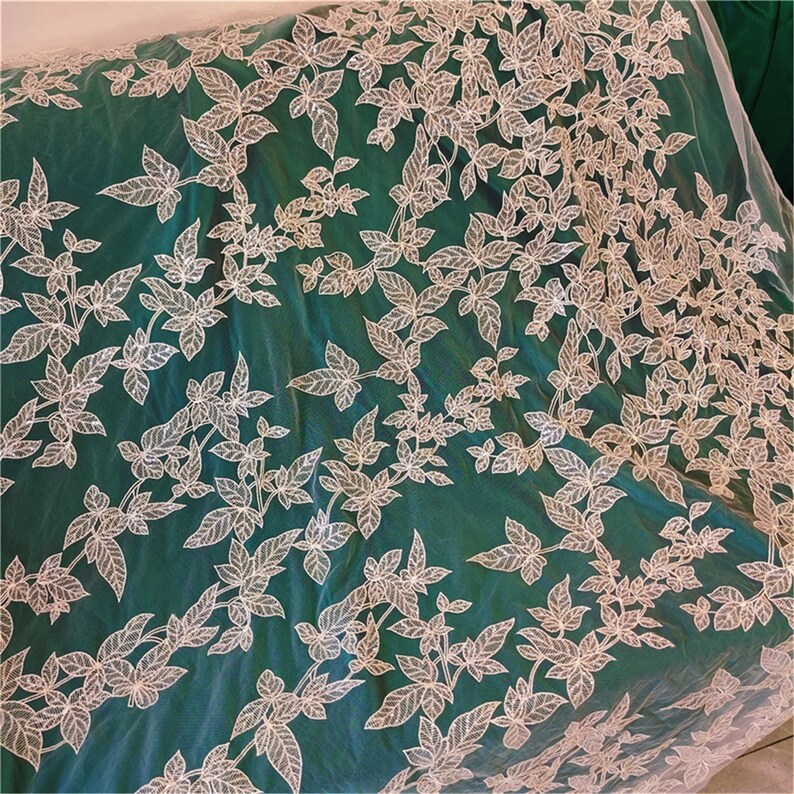 May include: White floral lace fabric with a green background. The lace has a delicate leaf pattern.
