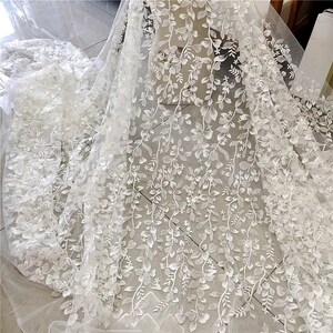 May include: White floral lace fabric with a delicate vine pattern. The lace is sheer and has a soft, flowing texture.