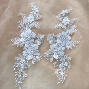 以下が含まれることがあります： 複雑な刺繍、透明なスパンコール、小さな白いビーズが施された2つの白い花のアップリケ。重なり合う花びら、葉のついた小枝、繊細な蔓のような巻きひげで構成され、左右対称の細長い植物模様が描かれています。